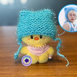 May include: A plush yellow chick doll wearing a light blue knitted cat ear hat, a pink scarf, and a pearl necklace. A smaller image shows a person wearing a similar hat. The doll has a logo on its base.