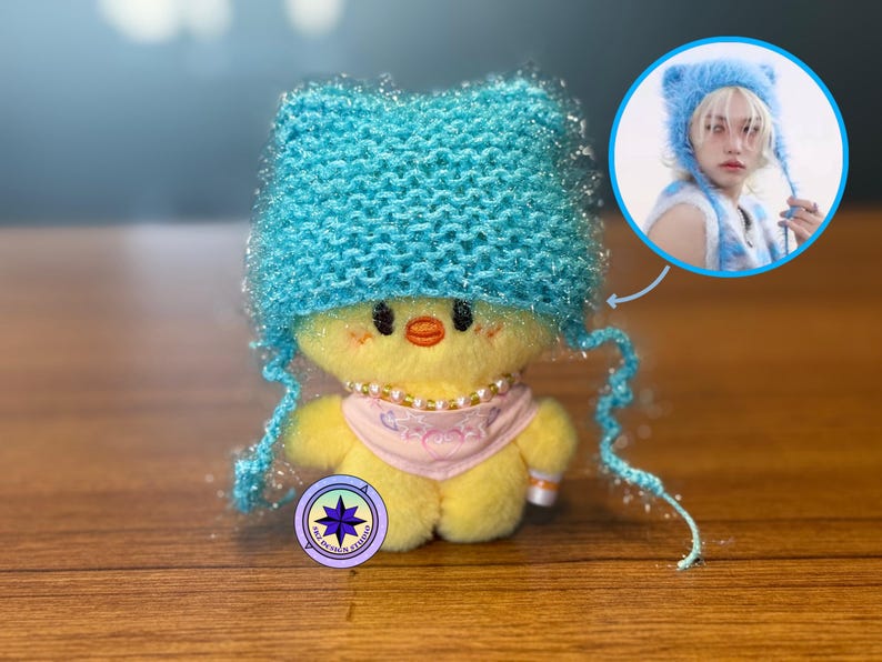 May include: A plush yellow chick doll wearing a light blue knitted cat ear hat, a pink scarf, and a pearl necklace. A smaller image shows a person wearing a similar hat. The doll has a logo on its base.
