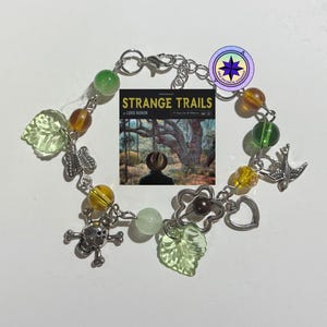 Può includere: Un braccialetto con ciondoli in argento con ciondoli verdi, marroni e argentati. I ciondoli includono un teschio, una farfalla, un uccello, un cuore, una foglia e un libro intitolato "Strange Trails" di Lord Huron.