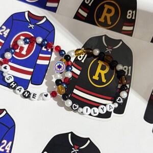 May include: A beaded bracelet with the name "SHANE" spelled out in white letter beads, and another with "OLYA". The bracelets are displayed with hockey jersey stickers in blue and black, with the numbers 24 and 81.