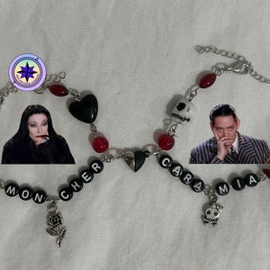 May include: A black and silver charm bracelet with red beads, black hearts, and skull charms. The bracelet has the words "Mon Cher" and "Cara Mia" spelled out with black letter beads.