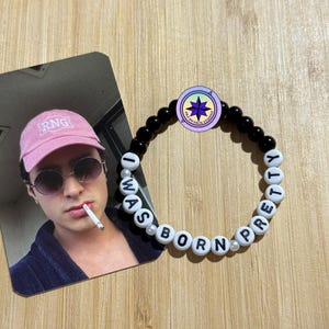 May include: A beaded bracelet with the words "I WAS BORN PRETTY" spelled out in white letter beads. The bracelet features black beads and a circular charm with a star design. A photo card of a person wearing a pink cap and sunglasses is next to the bracelet.