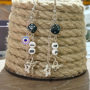 May include: Dangle earrings with black dice, white number beads, and small charms. The earrings feature a dice bead, followed by beads with the numbers 18 and 24. Charms include keys, hearts, and small pearl-like beads.