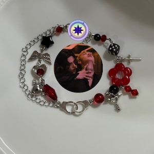 Pode incluir: Um bracelete de prata com contas vermelhas e pretas, os pingentes incluem um coração, asas, uma flor, um dado, algemas e uma chave. O bracelete também apresenta uma pequena imagem de um casal.
