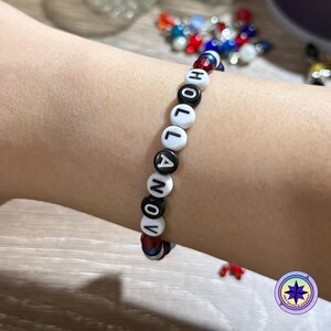 Op de afbeelding: Een armband met witte en zwarte letterkralen met de tekst "HOLLANOV". De armband is geregen op een rood, wit en blauw koord. De armband wordt om een pols gedragen.