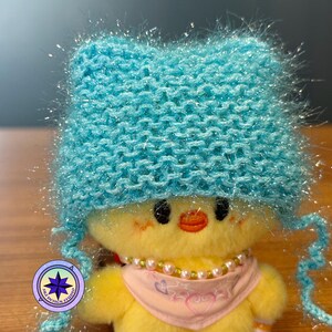 May include: A plush yellow chick wearing a light blue knitted hat with ear flaps and a pink scarf adorned with pearls. The chick has an orange beak and is set against a blurred background.