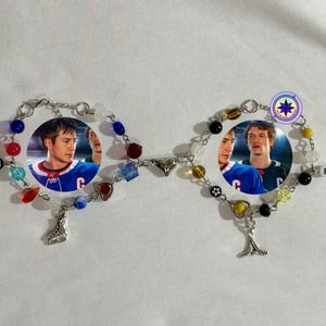 May include: Two beaded bracelets connected by a chain. Each bracelet features a circular image of a person, surrounded by colorful beads, charms, and letters. One bracelet has the letters "SH", the other "OR".