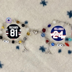 May include: Two beaded bracelets with hockey-themed charms. One bracelet features a black and white jersey design with the name "Rozanov" and the number "81". The other bracelet has a blue and white jersey design with the name "Hollander" and the number "24".