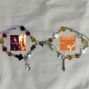 May include: Two beaded bracelets connected by a heart-shaped charm. One bracelet features a black cross charm, a guitar charm, and a My Chemical Romance album cover. The other has a microphone charm, a cross charm, and a The Smiths album cover.