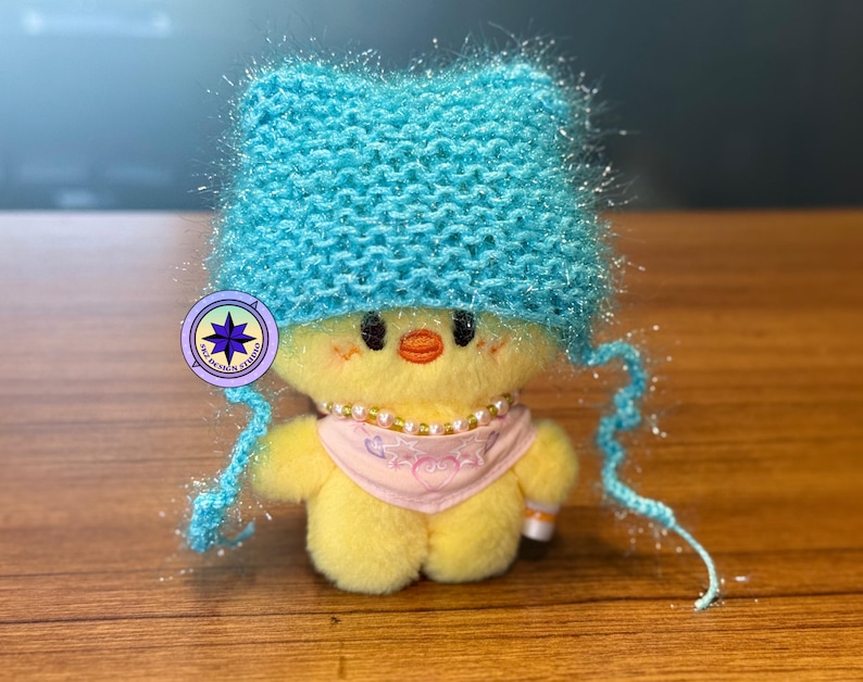May include: A small yellow plush chick wearing a bright turquoise knitted hat with long dangling ties, a beaded necklace, and a light pink bandana featuring a heart and star graphic. A circular logo with a purple star and text is attached.