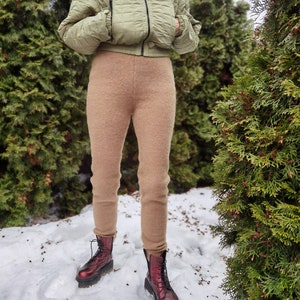 Op de afbeelding: Een vrouw draagt een lichtgroen gewatteerd jack en een bruine fleecebroek. Ze draagt rode leren laarzen met zwarte veters. De vrouw staat in de sneeuw.