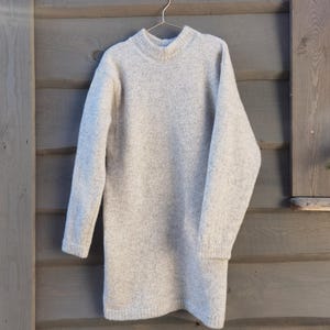 Peut inclure: Une robe pull grise clair à manches longues avec un col rond, suspendue à un cintre en métal. La robe est faite d'une matière douce et texturée. Le fond est un mur en bois avec des planches horizontales.