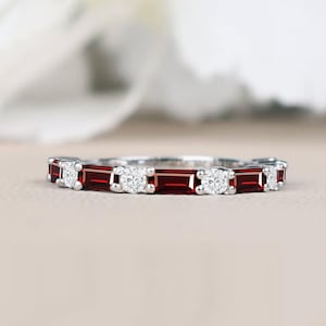 Puede incluir: Un anillo de banda en tono plateado que presenta un patrón alterno de piedras rojas de corte baguette y pequeñas piedras redondas transparentes de corte brillante, dispuestas en una fila continua sobre el frente para un diseño simétrico.