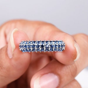 May include: A silver ring with a band of blue sapphires. The ring is being held by a hand.