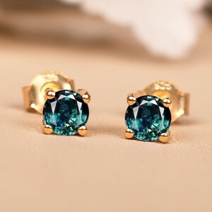 May include: A pair of gold stud earrings, each with a round, teal gemstone. The earrings are set on a neutral background, with soft, blurred white floral elements in the upper left corner.