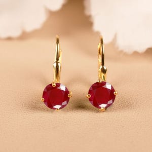 Può includere: Un paio di orecchini a leva color oro, ciascuno con una gemma rotonda di colore rosso intenso. Gli orecchini sono ambientati su uno sfondo neutro, con morbidi accenti floreali bianchi nella parte superiore dell'immagine.