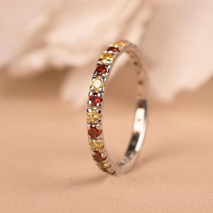 Könnte beinhalten: Ein silberner Ring mit einem Band aus abwechselnd roten, gelben und orangen Edelsteinen. Der Ring ist vor einem weichen, neutralen Hintergrund mit einer verschwommenen weißen Blume platziert. Es ist ein Schmuckstück.