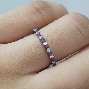 May include: A delicate ring featuring alternating red and clear gemstones set in a thin band. The ring is shown on a finger, showcasing the sparkle of the gemstones. The ring's design is simple yet elegant, suitable for everyday wear.