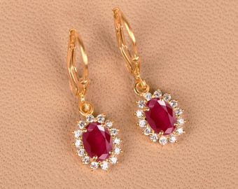 May include: A pair of gold-toned drop earrings featuring oval-cut deep red stones at the center, each surrounded by a halo of small, clear, sparkling stones. The earrings are attached to gold-toned lever-back closures for secure fastening.
