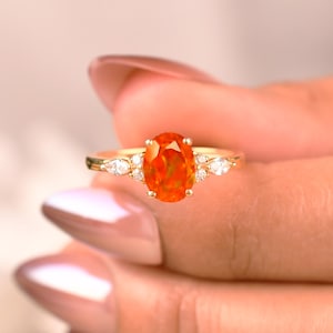 May include: A gold ring featuring an oval-cut orange gemstone as the centerpiece. The ring is accented with smaller, clear gemstones on either side of the main stone, set on a delicate band. The ring is a piece of jewelry.
