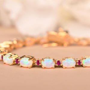 Op de afbeelding: Goudkleurige armband met ovale opalen en kleine rode edelstenen. De opalen hebben een melkachtige witte uitstraling met een subtiel kleurenspel. De armband is gezet op een gouden ketting en sluiting.