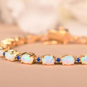 Op de afbeelding: Een gouden armband met ovale opalen en kleine blauwe edelstenen. De opalen zijn gezet in individuele gouden monturen, afgewisseld met blauwe stenen. De sluiting is ook van goud.