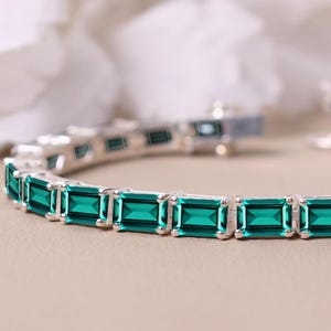 May include: A silver bracelet featuring rectangular emerald-green gemstones. The gemstones are set in a row, creating a continuous pattern. The bracelet is displayed on a neutral-toned surface, with a blurred background.