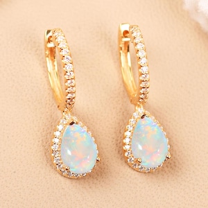 Puede incluir: Un par de pendientes de aro de oro con una piedra de ópalo en forma de lágrima engastada en un halo de circonita cúbica transparente. Los pendientes tienen un acabado dorado pulido.