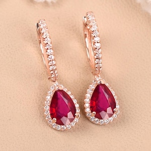 Puede incluir: Un par de pendientes de oro rosa con una piedra preciosa de rubí en forma de lágrima rodeada por un halo de piedras preciosas más pequeñas y transparentes. Los pendientes tienen un diseño de aro.