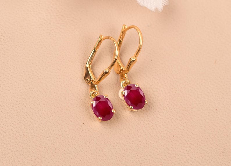 May include: A pair of gold earrings with red oval-shaped gemstones. The earrings have a lever back closure.