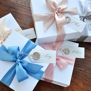 May include: Four white gift boxes with different colored ribbons and personalized tags. The boxes are tied with a light blue, pink, cream, and gray ribbon. Each tag has a wax seal and a name written in calligraphy. The names on the tags are "Jenna", "Avery", "Rebecca", and "Julie".