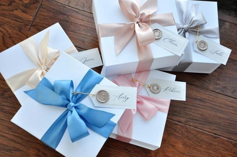 May include: Four white gift boxes with different colored ribbons and personalized tags. The boxes are tied with a light blue, pink, cream, and gray ribbon. Each tag has a wax seal and a name written in calligraphy. The names on the tags are "Jenna", "Avery", "Rebecca", and "Julie".