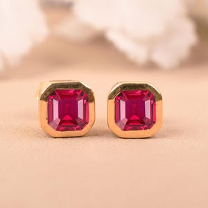 May include: A pair of gold-coloured square stud earrings featuring vibrant, square-cut ruby-red gemstones. The earrings are set in a gold bezel setting, creating a classic and elegant look. The background is a soft, neutral colour.