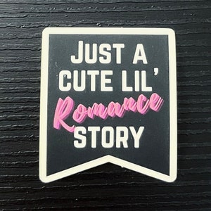 May include: A black and white rectangular sticker with the text "JUST A CUTE LIL' Romance STORY" in white and pink lettering. The sticker has a banner shape with a white border.