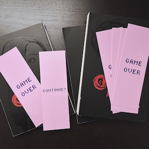 May include: Pink bookmarks with pixelated text that reads "GAME OVER" and "CONTINUE?" are placed near black books with red circular designs. The books have illustrations of a person's face. The bookmarks are rectangular.