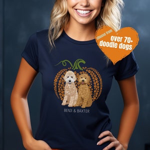 May include: A navy blue t-shirt with a pumpkin design featuring two doodle dogs. The pumpkin is made of paw prints and the text "Benji & Baxter" is printed below the dogs. The text "choose from over 70+ doodle dogs" is printed on a heart-shaped orange tag.