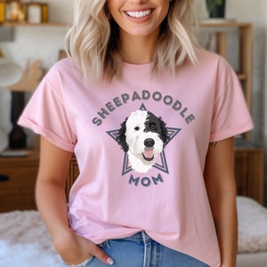May include: A pink t-shirt with a graphic of a black and white sheepadoodle dog inside a blue star with the text "Sheepadoodle Mom" in a retro font.