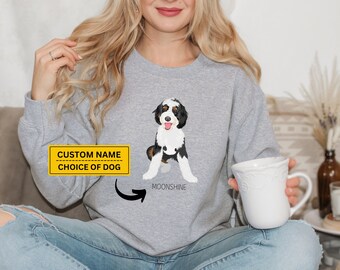 May include: A gray sweatshirt with a custom name and choice of dog printed on the front. The dog is a black, white, and brown doodle. The text "Moonshine" is printed below the dog.
