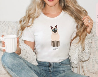May include: White t-shirt with a graphic of a Siamese cat sitting down. The cat is light brown with blue eyes and the text "OREO" is printed below the cat.