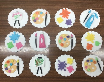 May include: Twelve round white cupcakes decorated with various art-themed fondant toppers. The toppers include paint palettes, paintbrushes, paint tubes, and canvases with flowers and trees.