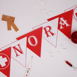 May include: A red and white banner with the word "NORA" spelled out in white letters on red triangles. The banner is decorated with red crosses on white circles. There is a red apple, a thermometer, and a syringe with red liquid in the image.