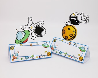 May include: Two white triangular place cards with a blue border. The place cards are decorated with a space theme, including a rocket, a telescope, a planet, a UFO, and stars. The place cards are blank for writing names.