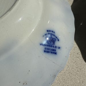 Puede incluir: Un plato de porcelana blanco con borde festoneado y detalles azules. El plato presenta el texto "Royal Porcelain" y "Wedgwood & Co. England Montrose" en azul.