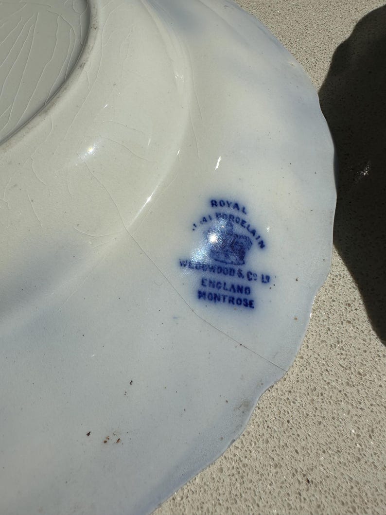 Puede incluir: Un plato de porcelana blanco con borde festoneado y detalles azules. El plato presenta el texto "Royal Porcelain" y "Wedgwood & Co. England Montrose" en azul.