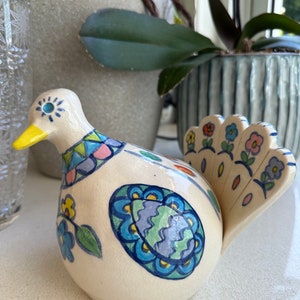 Puede incluir: Una figurita de pájaro de cerámica con una base blanca y detalles pintados de colores. El pájaro tiene un ojo azul, un pico amarillo y una cola con estampado azul, verde y amarillo.