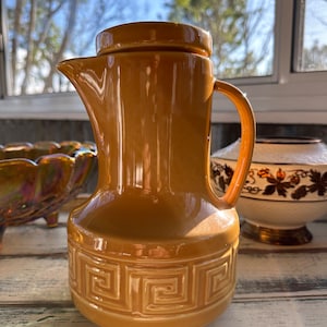 May include: A vintage, amber-colored ceramic pitcher with a lid and handle. The pitcher features a Greek key design around the base. The spout is pointed, and the handle is curved. The pitcher is likely for serving beverages.