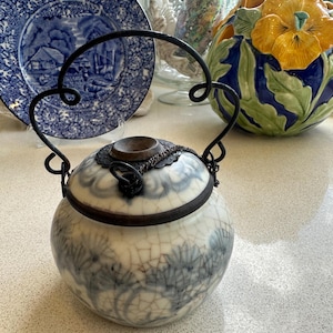 Puede incluir: Un tarro de cerámica blanca con un diseño floral azul y blanco y una tapa de metal. El tarro tiene un asa de metal y está sentado sobre una superficie blanca.