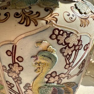 May include: A cream-colored ceramic vase with a detailed, raised relief design. The vase features a phoenix bird, floral patterns, and leaf accents in shades of green, brown, and gold. The vase has a crackled glaze and a wide, rounded shape.