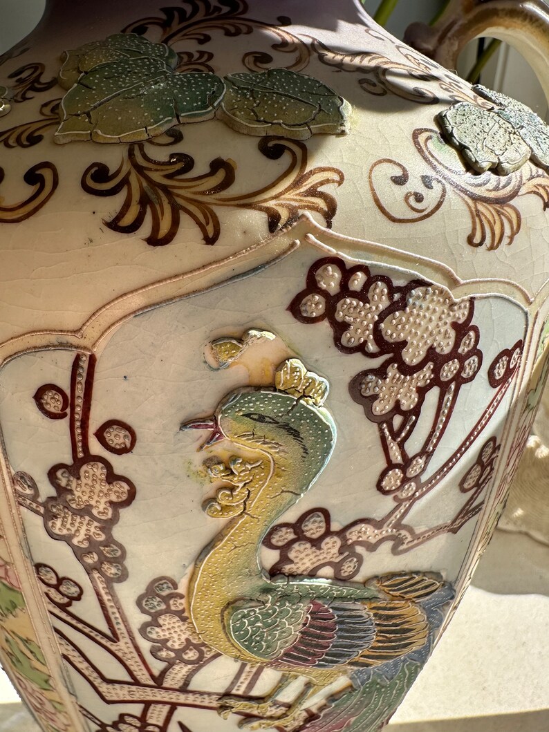 May include: A cream-colored ceramic vase with a detailed, raised relief design. The vase features a phoenix bird, floral patterns, and leaf accents in shades of green, brown, and gold. The vase has a crackled glaze and a wide, rounded shape.