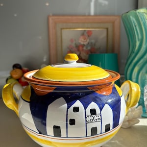 Puede incluir: Una sopera de cerámica con tapa, con un diseño pintado a mano de casas blancas sobre un fondo azul. La tapa es amarilla con detalles en naranja y blanco. La sopera tiene asas amarillas y un pomo a juego.